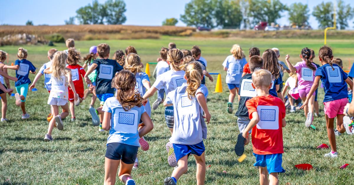  A large group of young kids running through a field. They've got signs on their back in support of a fundraising event.