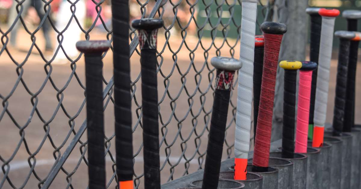 A row of metal baseball bats lined up along a chain link fence. The stickers and wraps around the handles are worn.