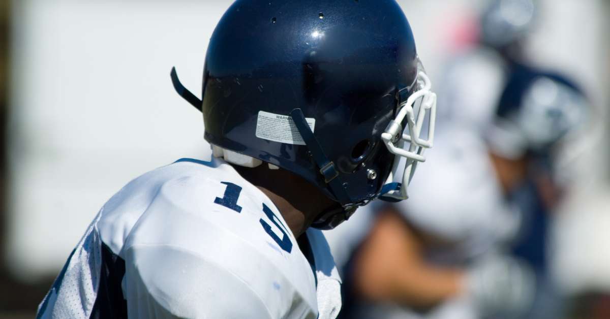 A young football player in a number 15 white and blue jersey. His helmet has a noticeable white sticker on it.